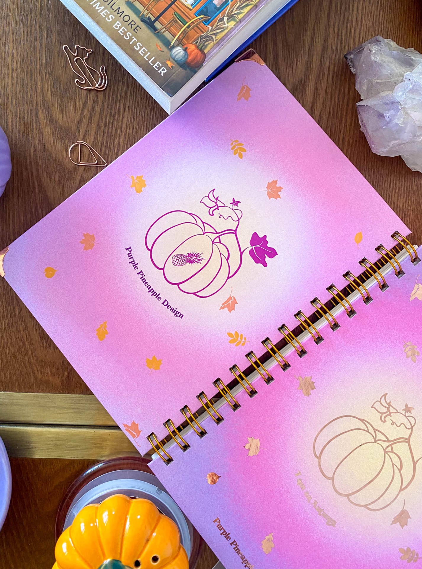 Notebook rosa degradê com ilustrações douradas de abóboras e folhas sobre mesa de madeira