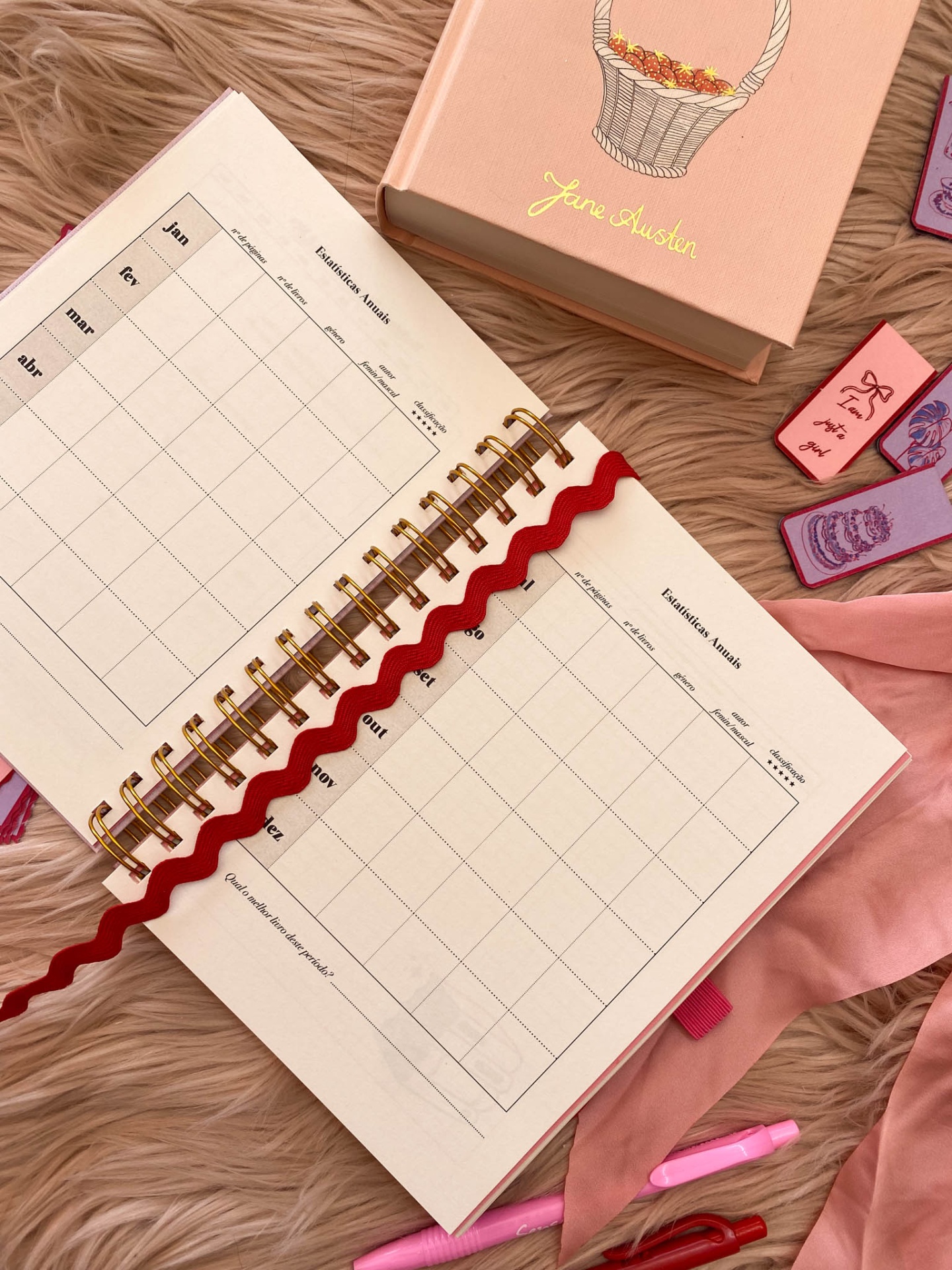 Caderno aberto com planejamento mensal, caixa rosa Jane Austen, carimbos e caneta sobre tapete peludo.