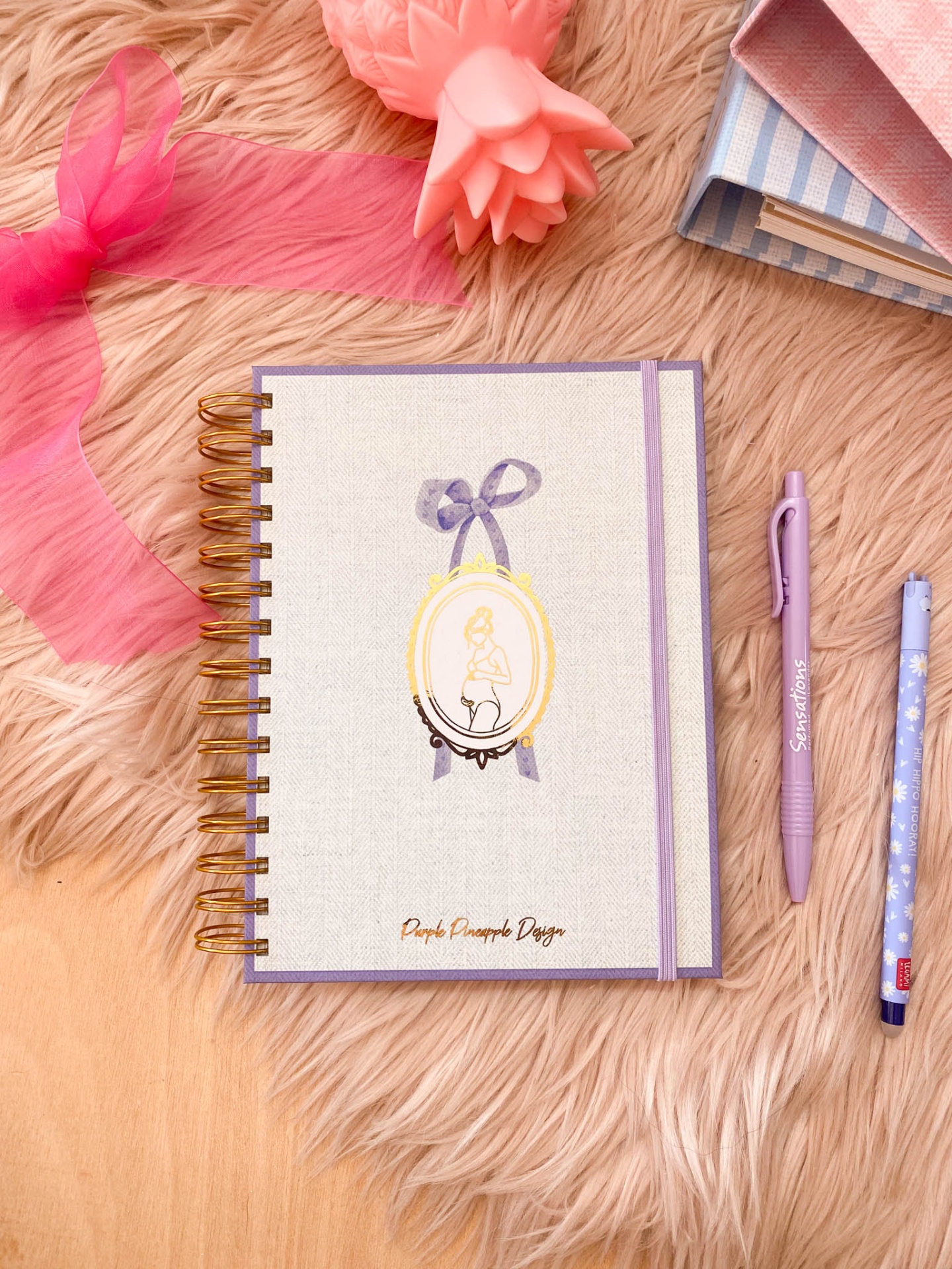 Caderno espiral branco e lilás com ilustração e texto, sobre pelo cor de creme com canetas, fita rosa e livros