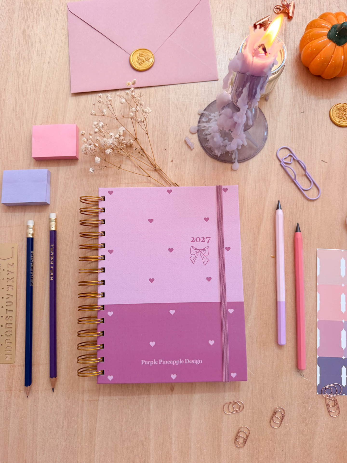 Planner 2027 rosa e roxo com selo de cera, lápis, canetas, clips e vela derretida sobre superfície de madeira