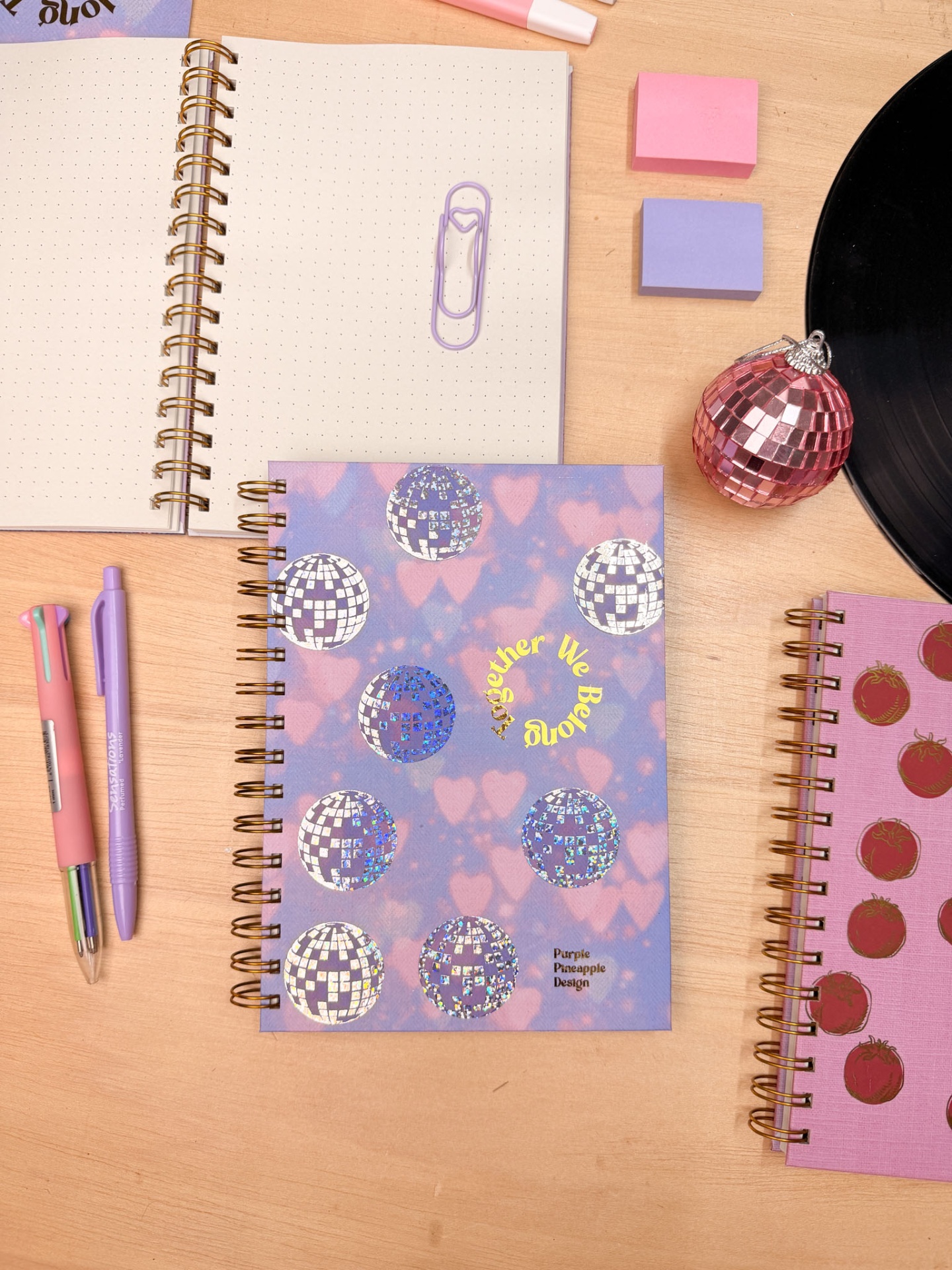 Caderno espiral azul e rosa com bolas disco e texto, sobre mesa com artigos de escrita e decoração.