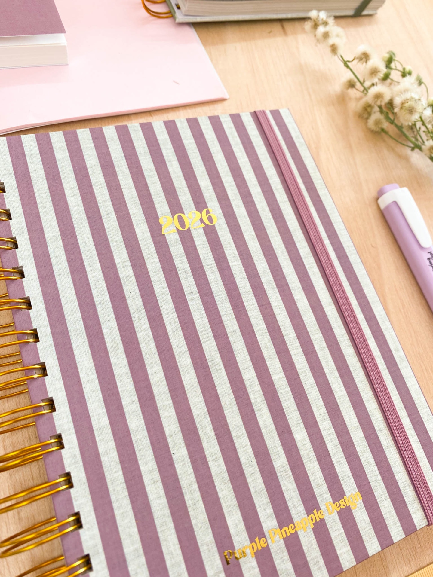 Agenda riscada lilás e branca com texto dourado, espiral dourada e elástico lilás numa mesa de madeira com caneta e flores