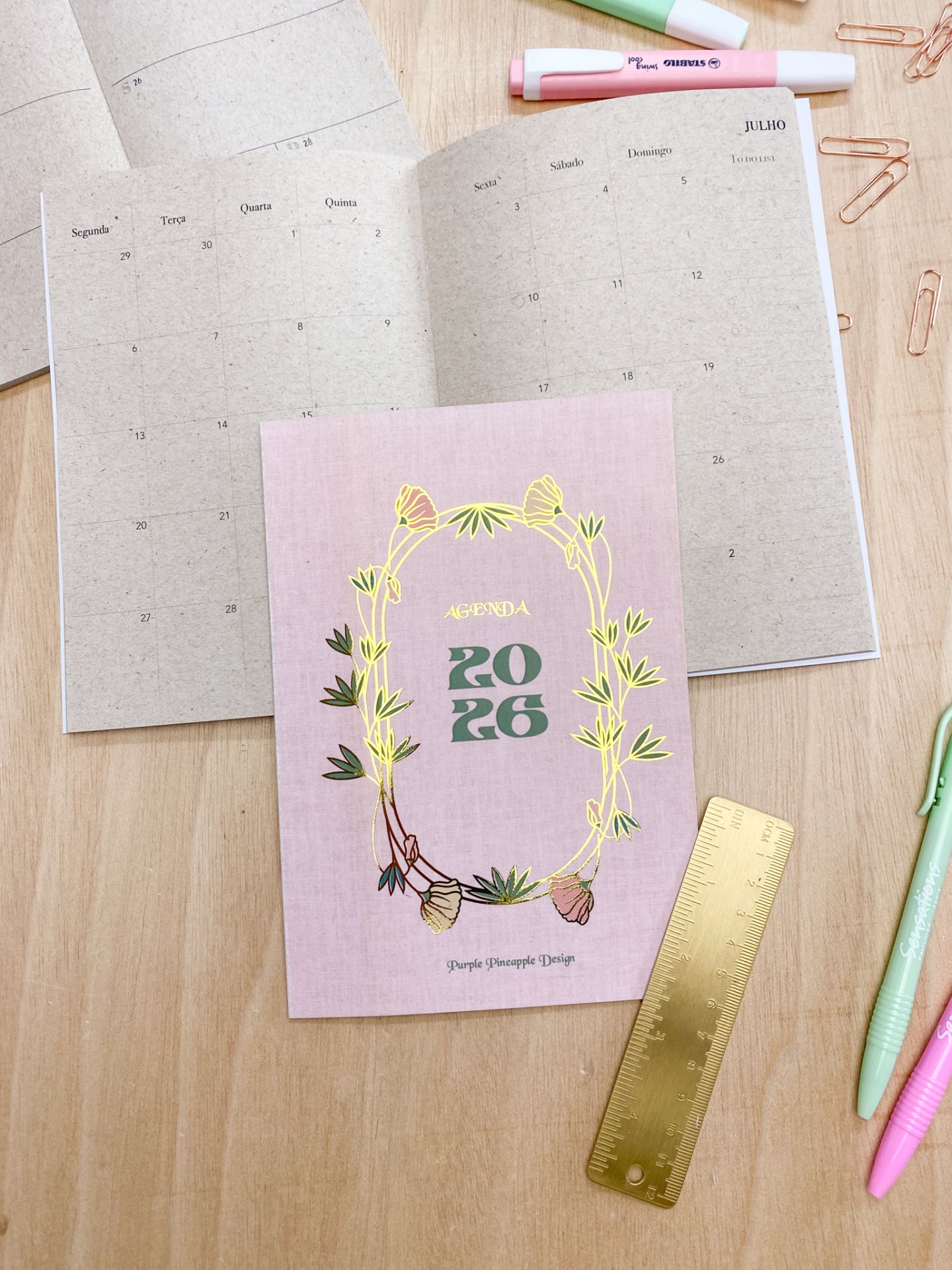 Agenda 2026 rosa com flores douradas sobre mesa com régua e canetas