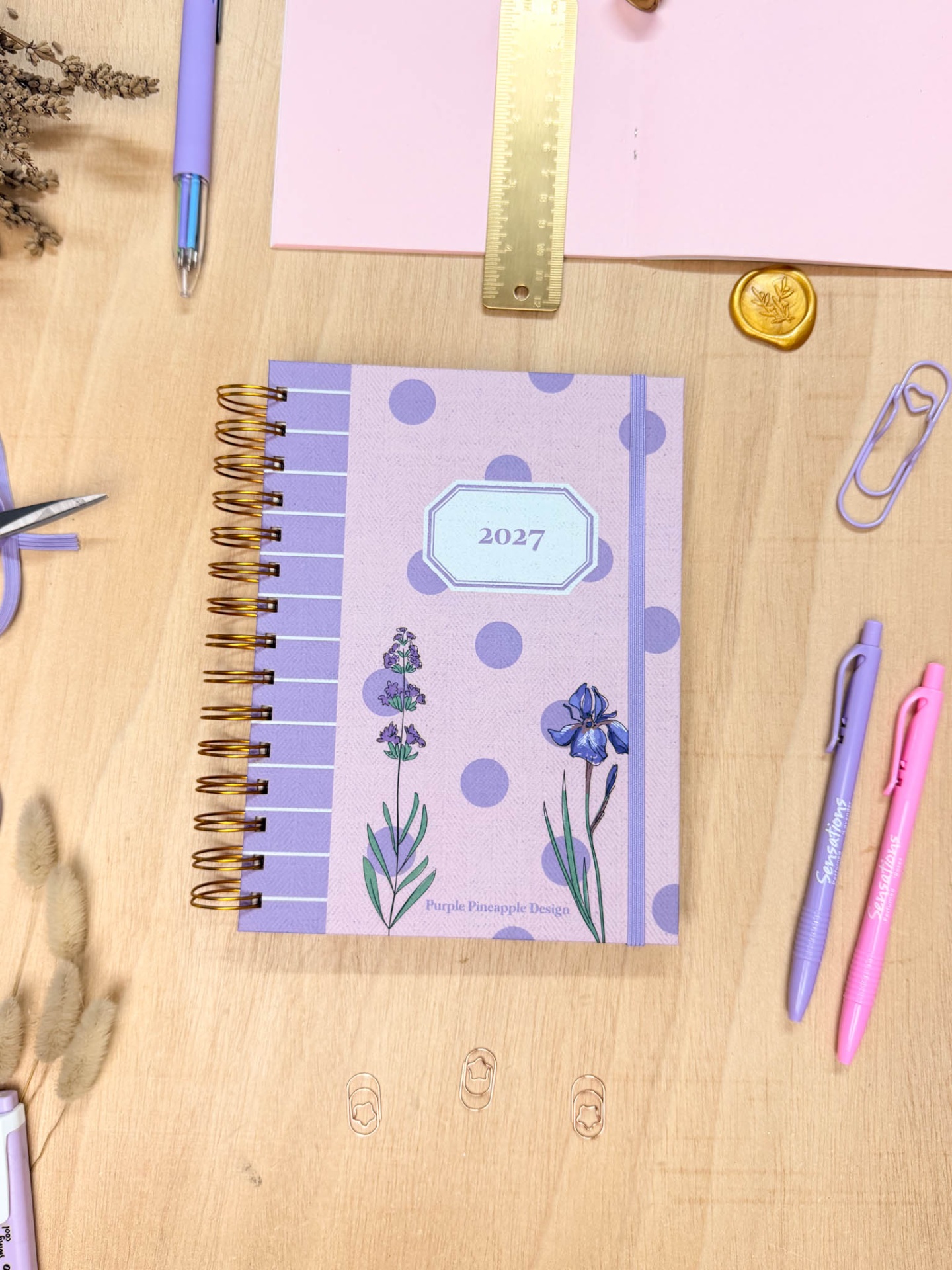 Caderno lilás com flores e bolinhas, texto 2027 e Purple Pineapple Design sobre mesa de madeira com material de escritório