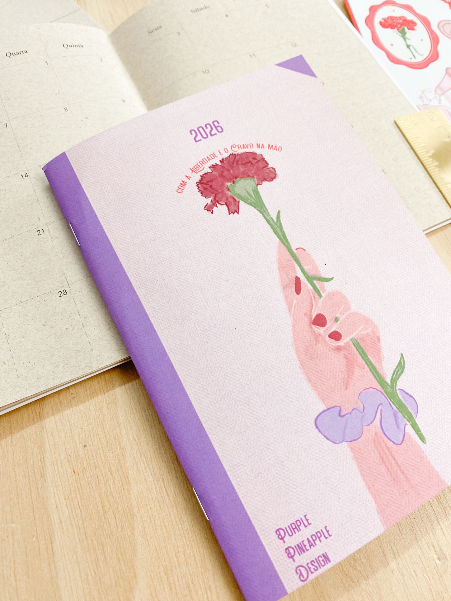 Agenda 2026 com ilustração de mão segurando cravo vermelho e texto em capa lilás e lombada roxa
