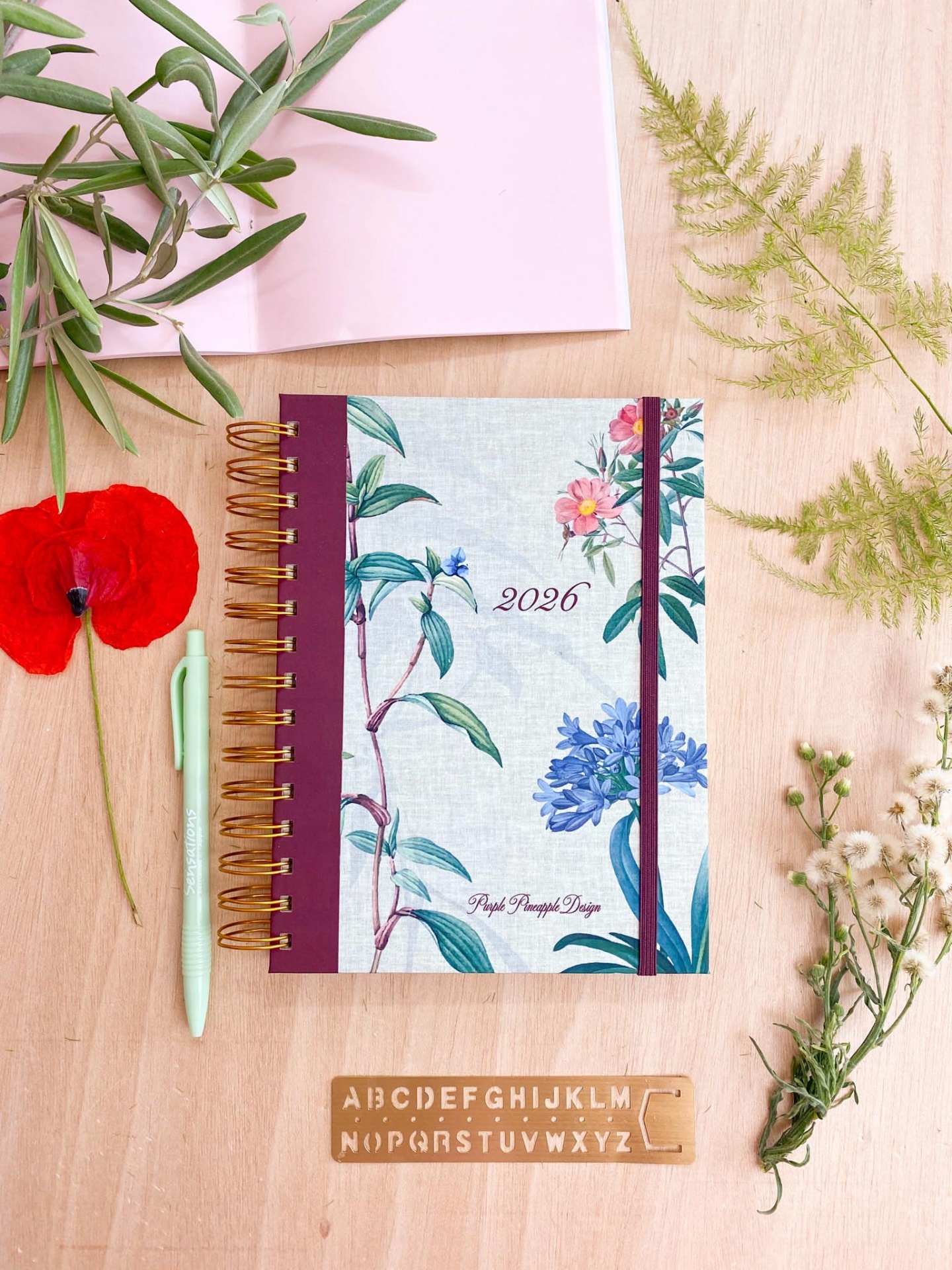 Agenda 2026 com capa floral, caneta e estêncil sobre mesa de madeira com flores