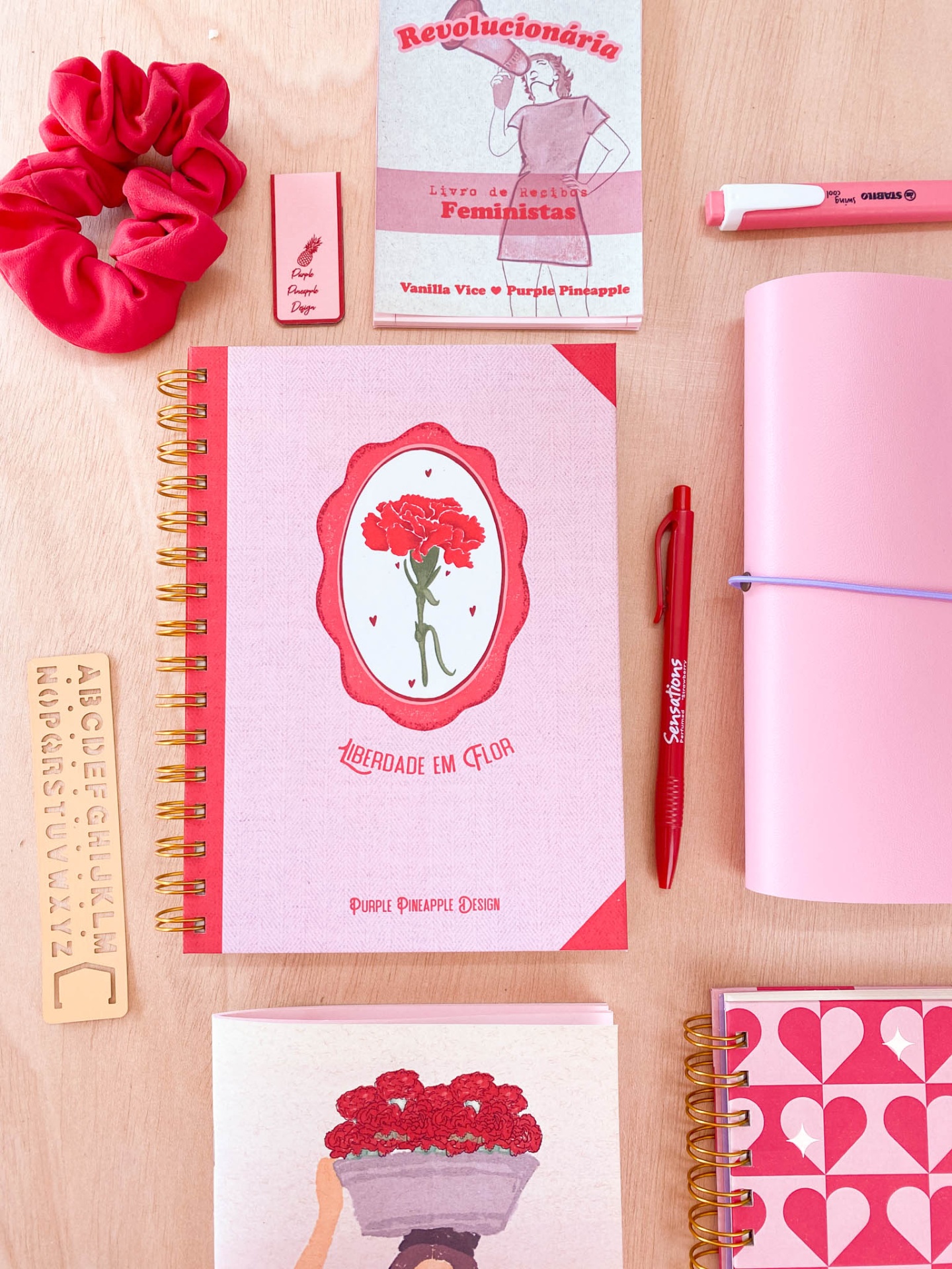 Material de papelaria rosa e vermelho com cadernos, canetas, régua de madeira e bloco de notas com palavra 'Revolucionária'