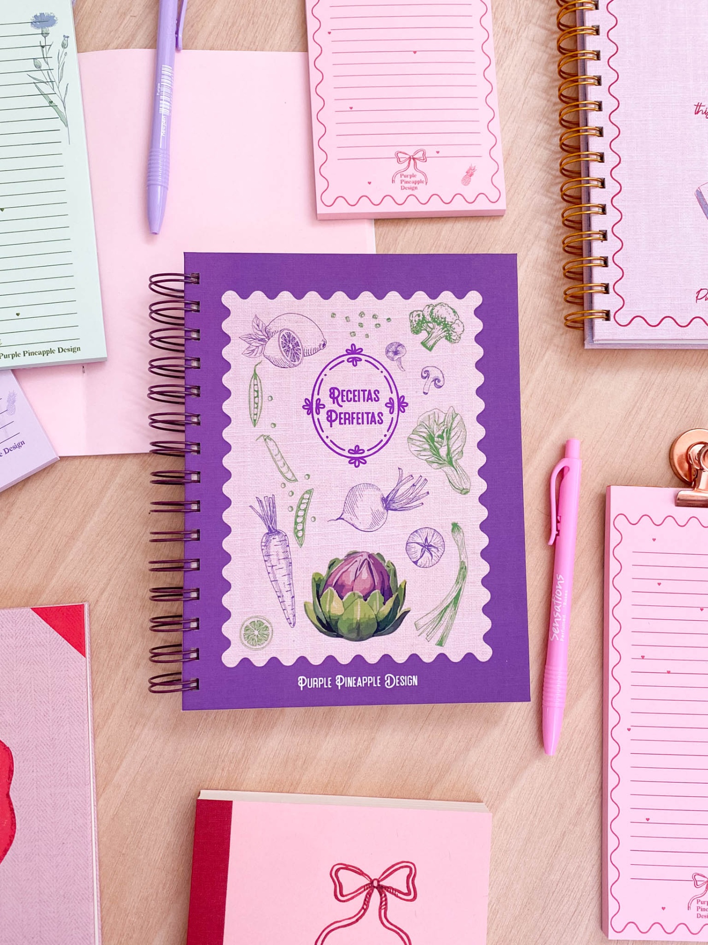 Caderno roxo e lilás com desenhos de legumes e texto RECETAS PERFEITAS rodeado por outros cadernos e canetas coloridas