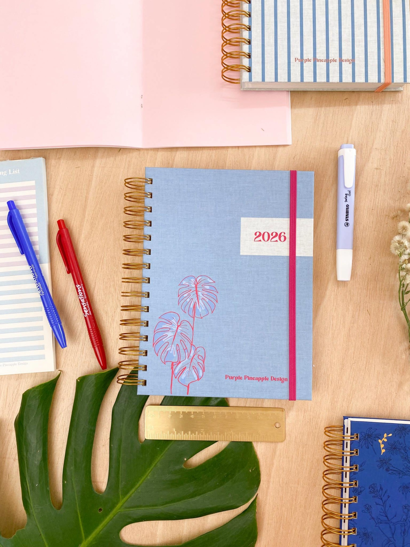 Cadernos de argolas douradas com capas azul, azul escuro e listrada, e inscrições '2026' e 'Purple Pineapple Designs'.