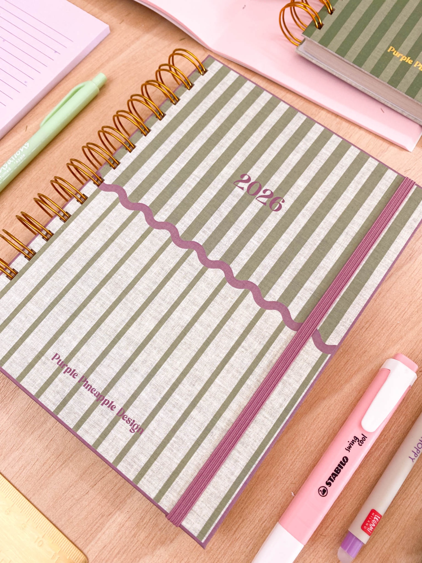 Caderno 2026 riscas cinza e branca com marcador púrpura e canetas numa mesa
