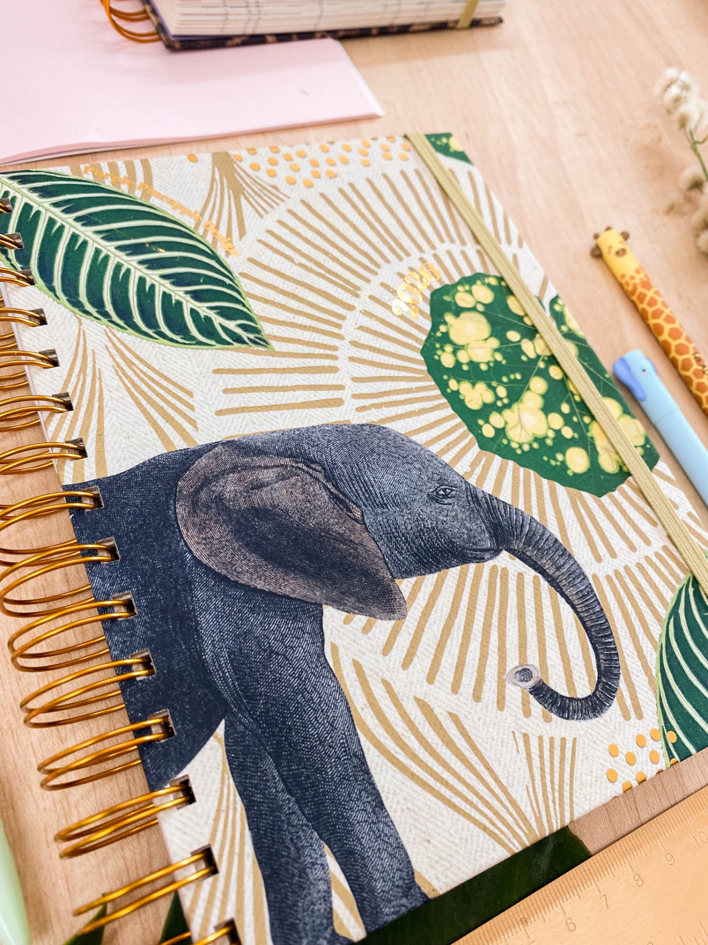 Caderno espiral com capa decorada com elefante e folhas, sobre mesa de madeira com canetas e régua