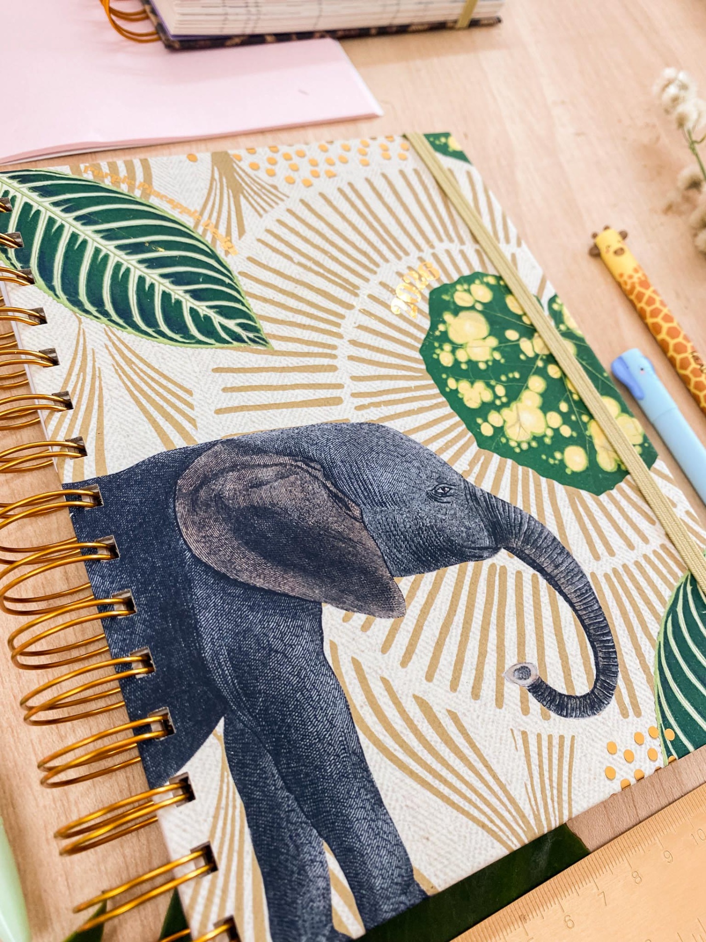 Caderno com capa de elefante e folhas decoradas, com canetas e mesa de madeira