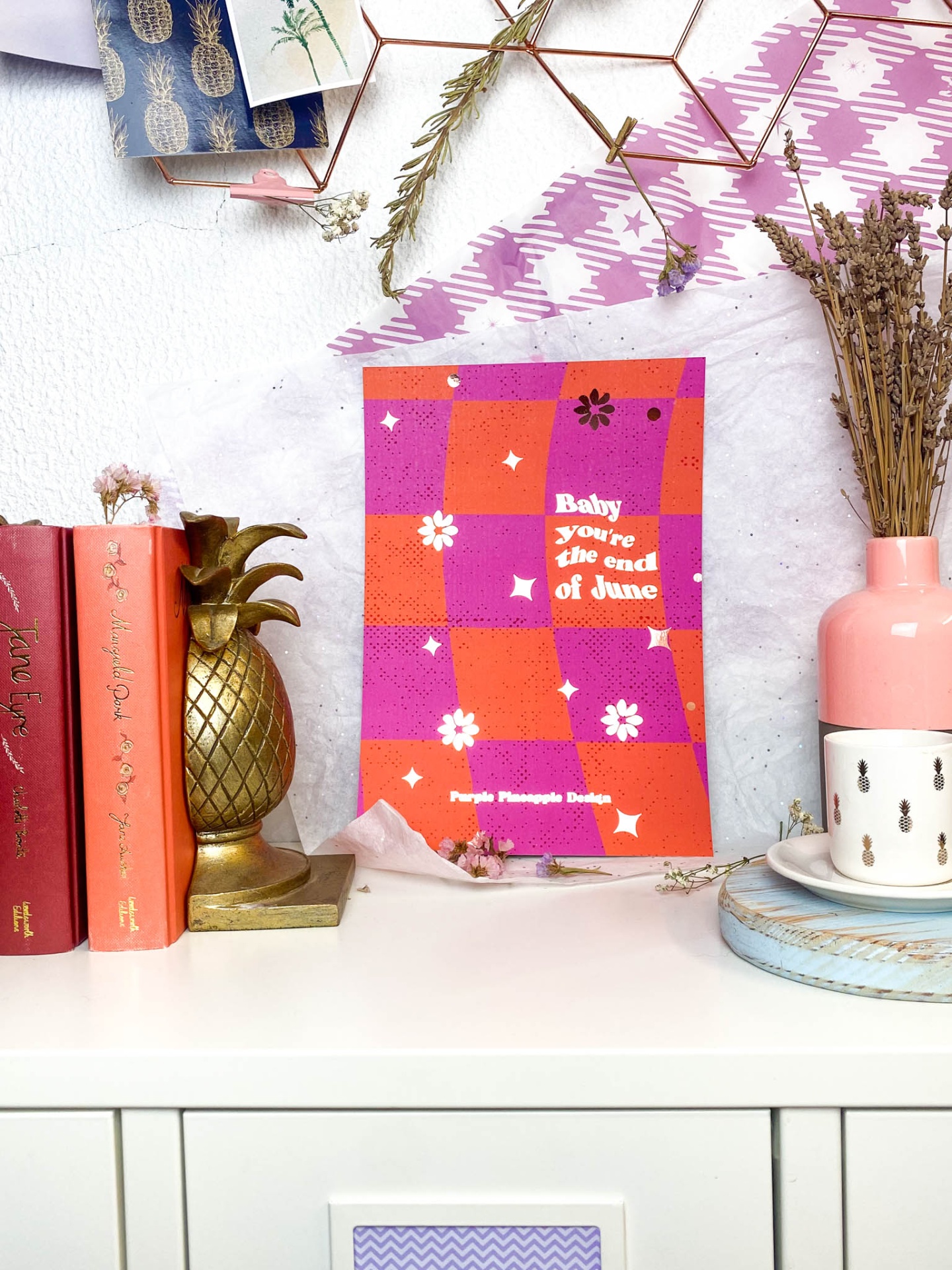 Cartaz roxo e laranja com texto branco em ambiente decorado com estatueta abacaxi dourada, livros vermelhos, flores secas num vaso rosa e chávena branca