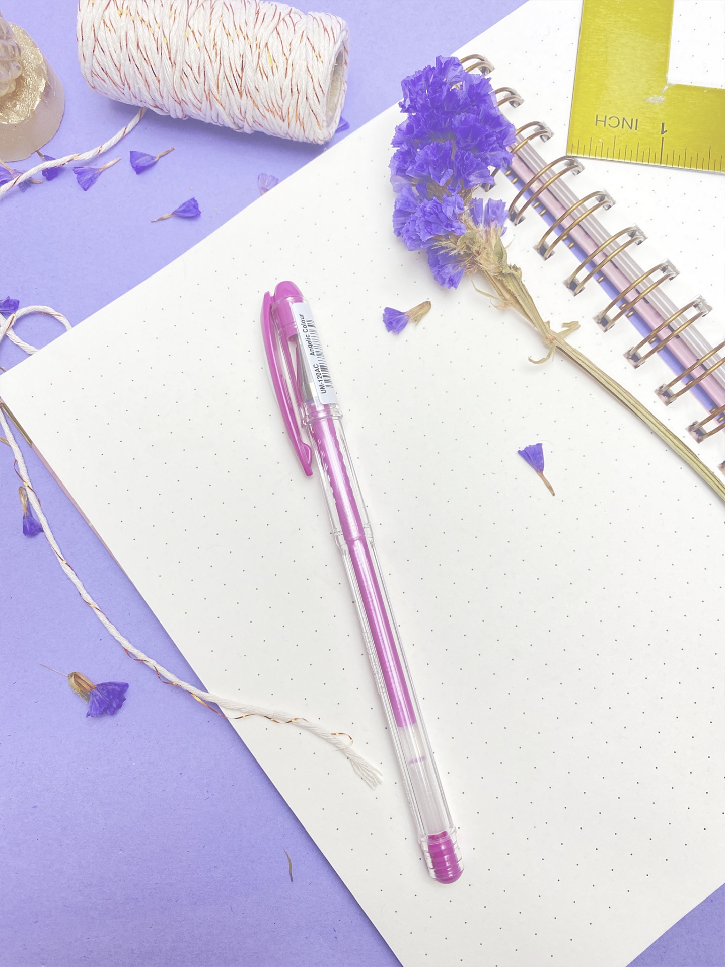 Caneta roxa, caderno pontilhado espiral, flores secas e cordel sobre superfície lilás