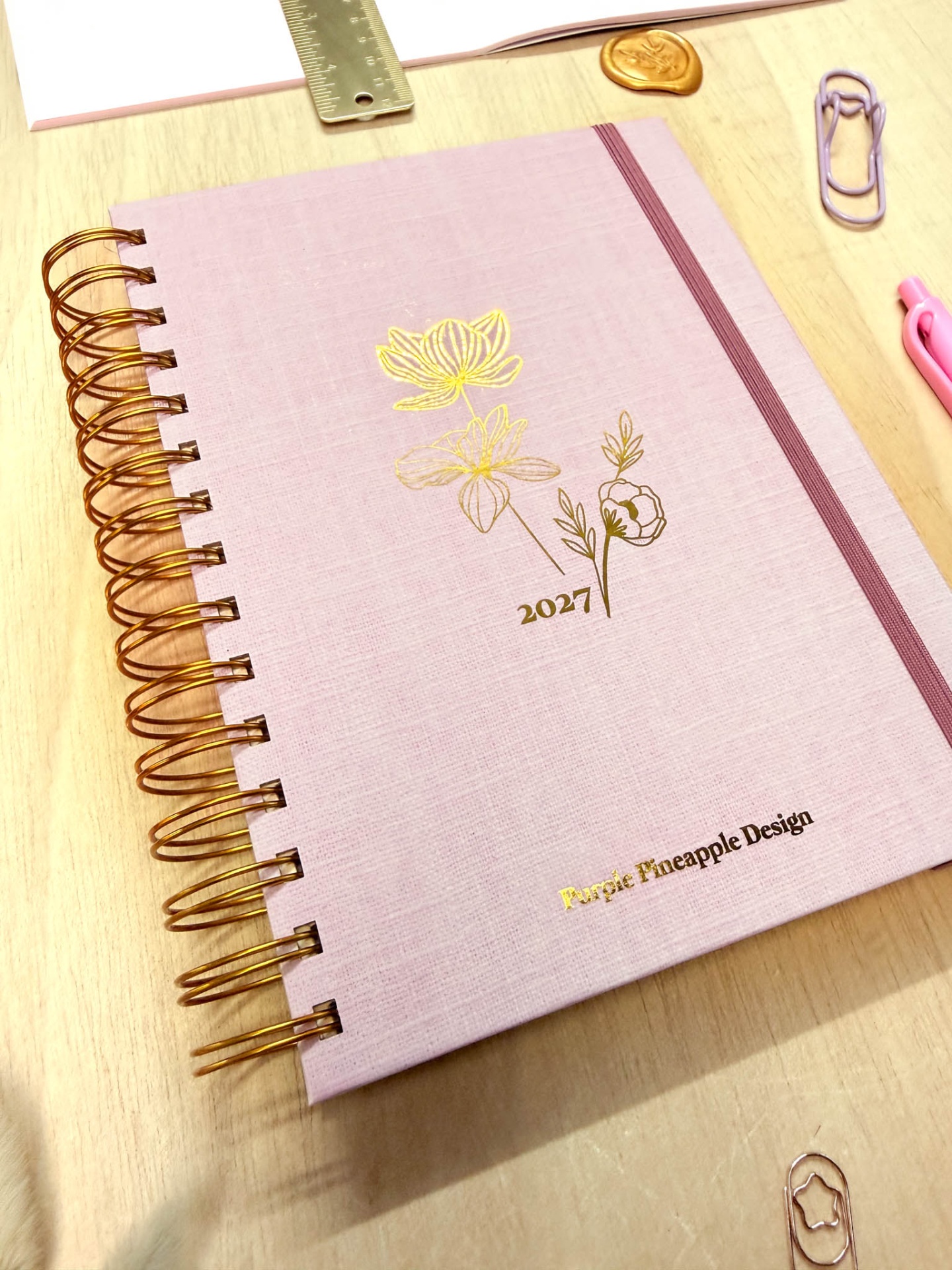 Agenda rosa claro com flores douradas e texto Purple Pineapple Design em mesa de madeira com acessórios de escritório.