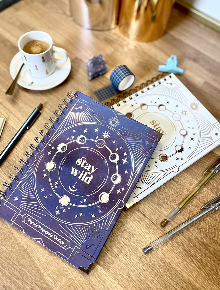 Dois cadernos espiralados com capas azul e branca com padrões astrológicos e texto 'stay wild' numa mesa de madeira com café e acessórios de escrita