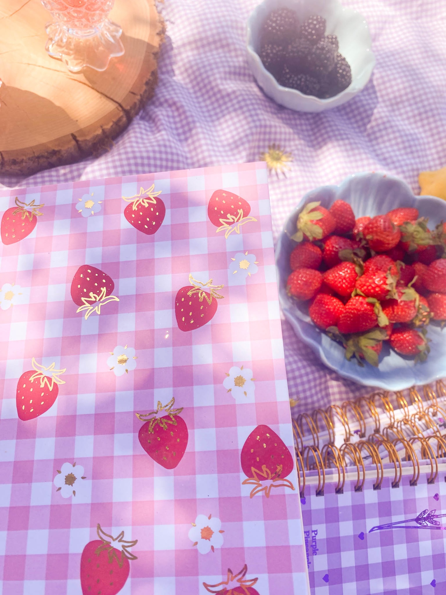 Caderno rosa com estampa de morangos e flores ao lado de pratos com morangos e amoras sobre tecido xadrez lilás.