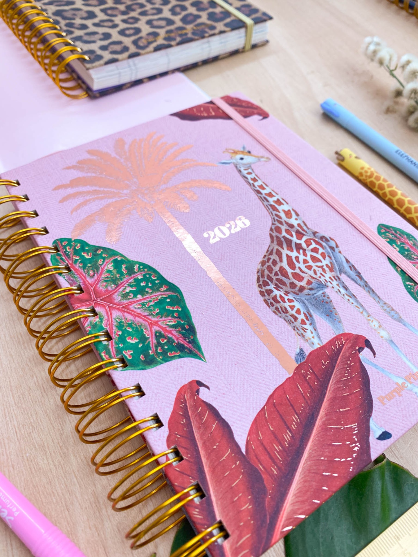 Caderno rosa com ilustrações de girafa e folhas tropicais e espiral dourada