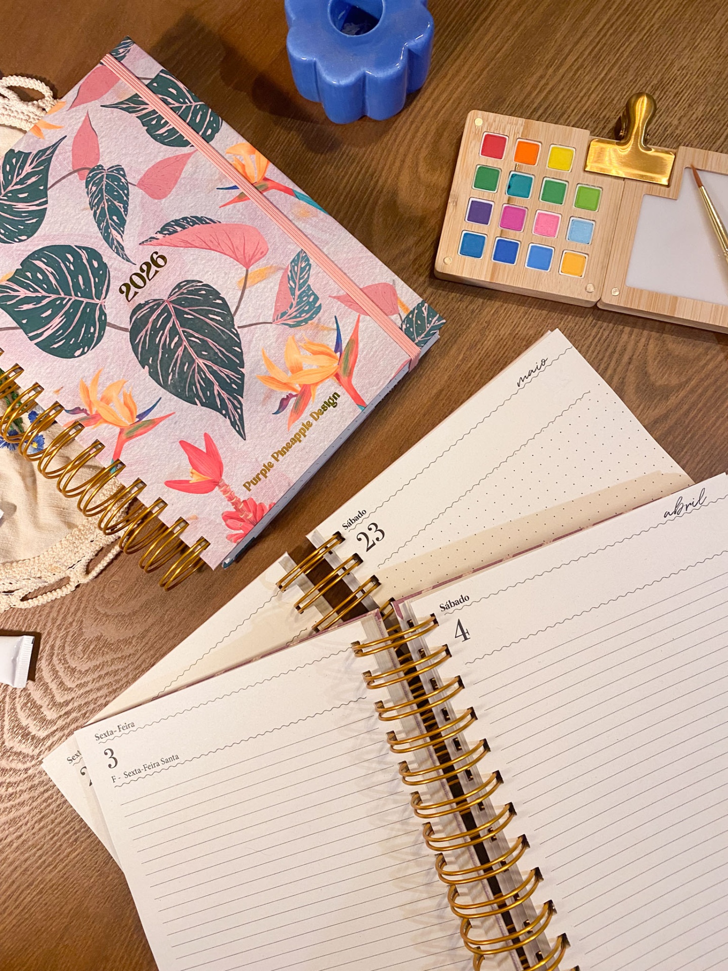 Agenda floral 2026 com páginas abertas, paleta de tintas coloridas e vela azul sobre mesa de madeira