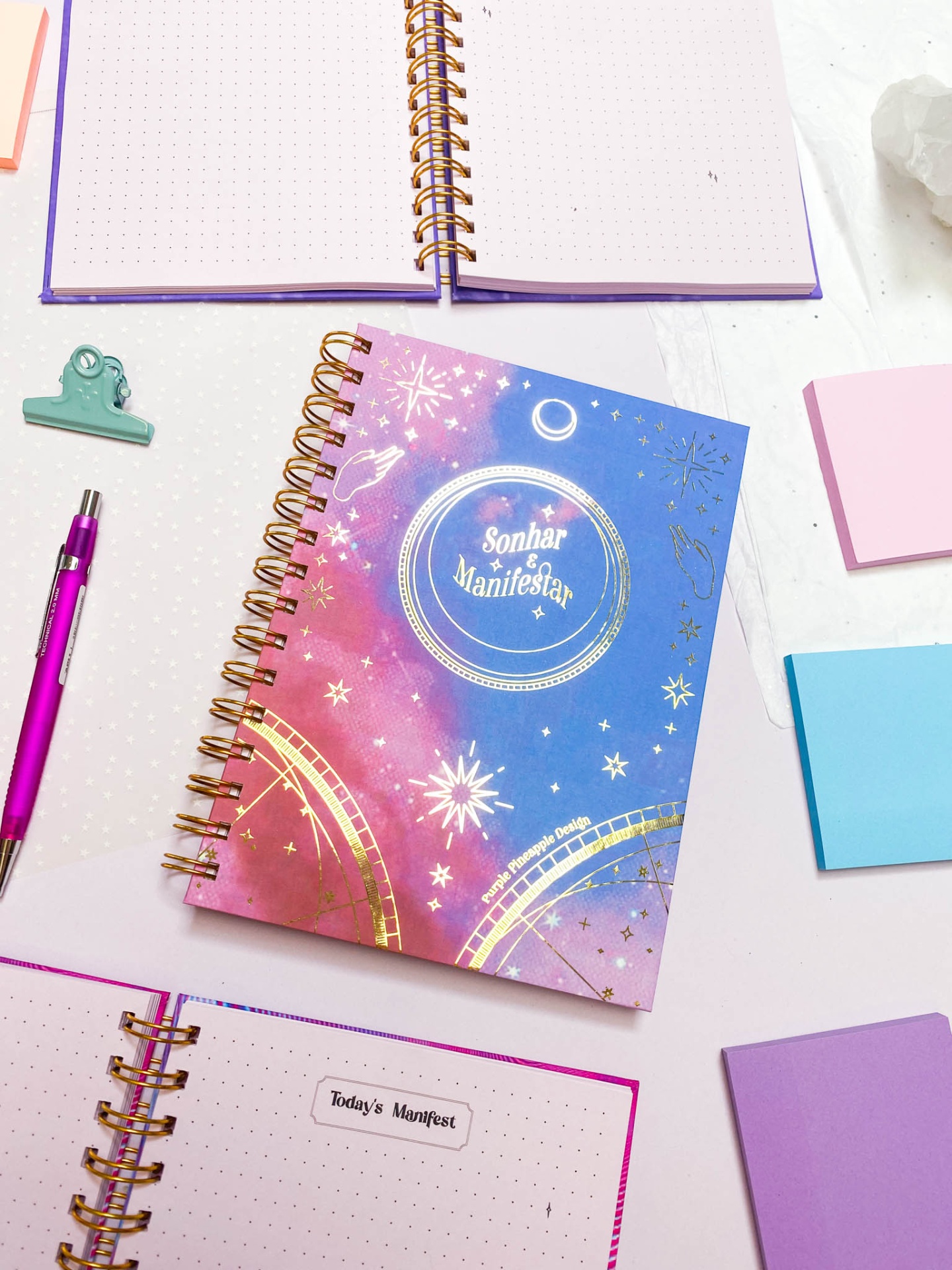 Caderno com capa azul e rosa com texto Sonhar é Manifestar e padrões cósmicos dourados