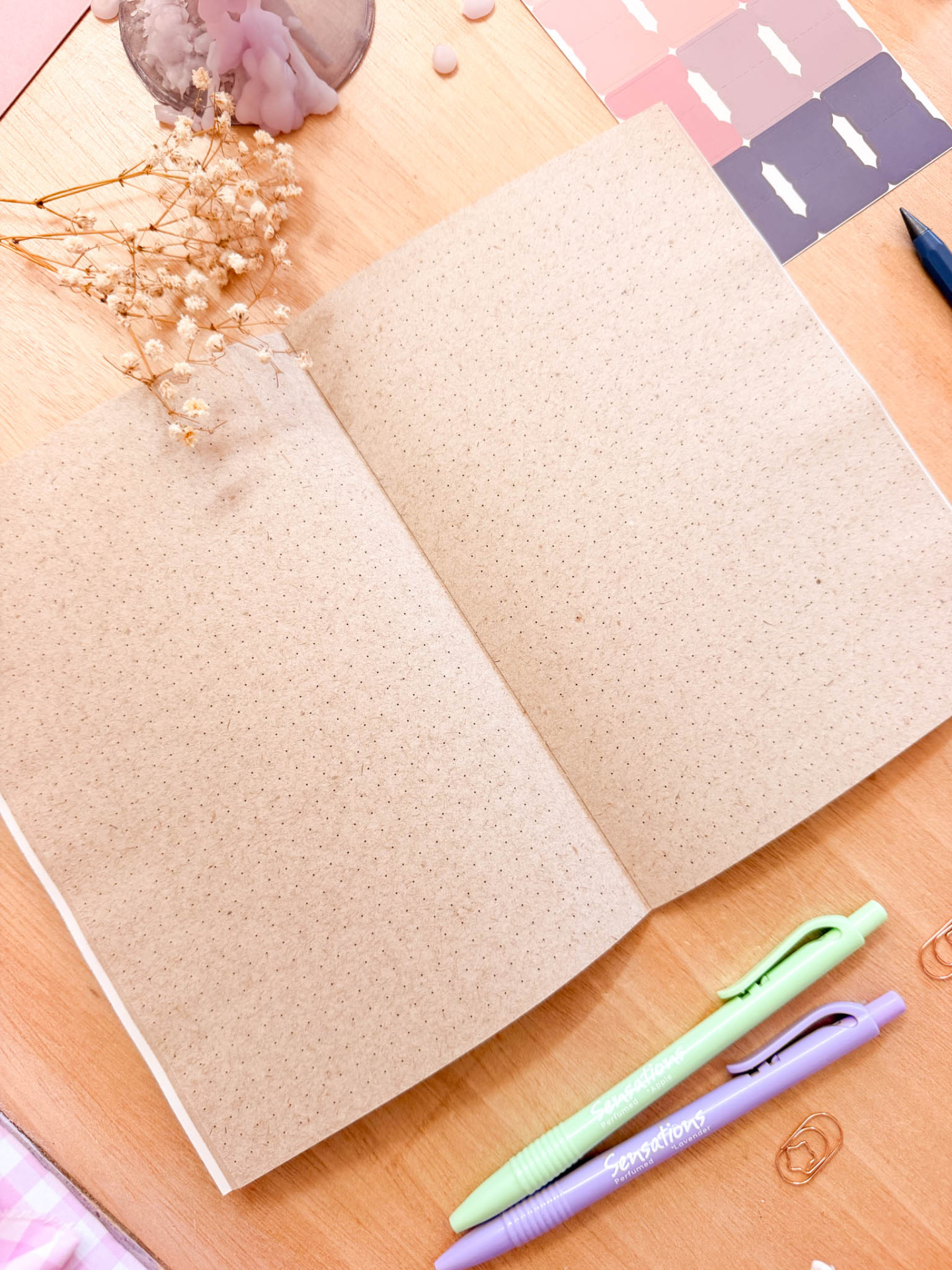 Caderno aberto em papel castanho pontilhado com canetas verdes e lilás Sensations e elementos de papelaria numa mesa de madeira