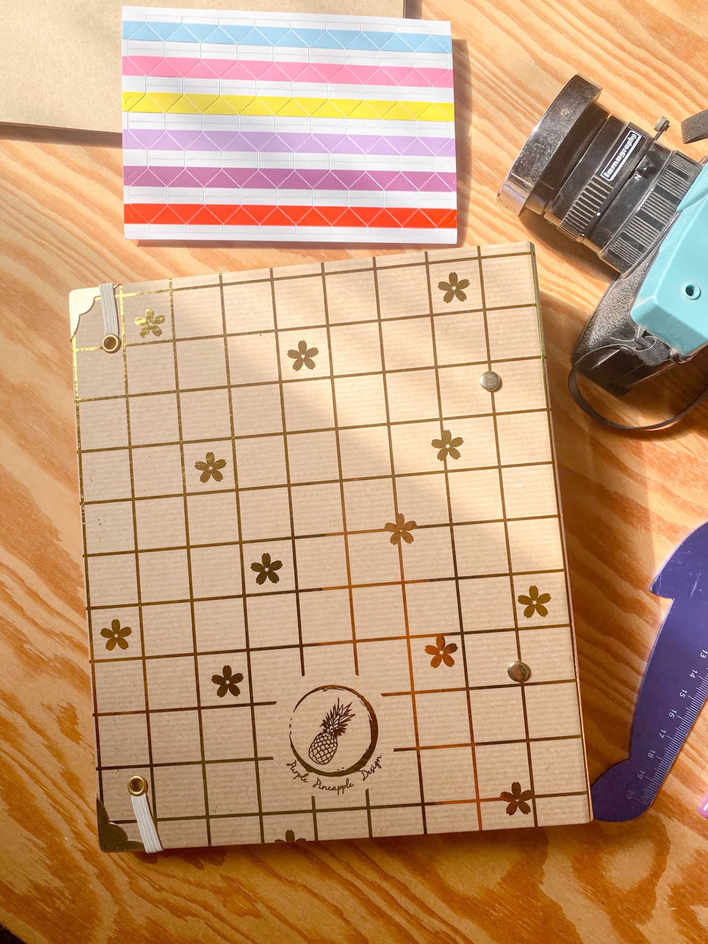 Caderno com capa em papel kraft decorada com flores e grelha, selo com abacaxi e texto THE PINEAPPLE PROJECT, sobre mesa de madeira com acessórios perto