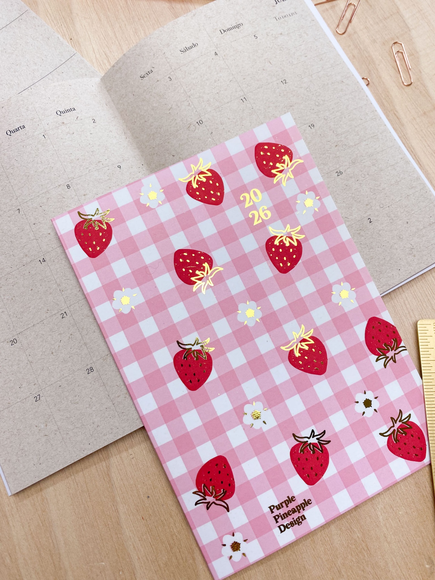 Agenda 2026 com capa em padrão xadrez rosa e branco com morangos e flores, sobre caderno bege e mesa de madeira.