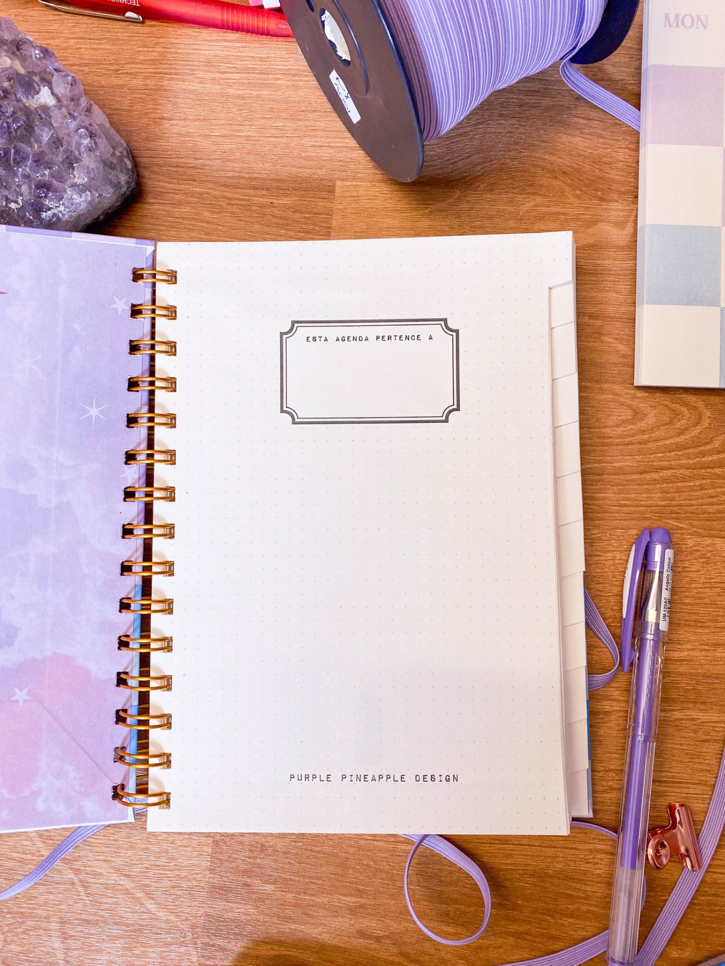 Caderno aberto com texto 'ESTA AGENDA PERTENCE A' e acessórios lilás sobre mesa de madeira