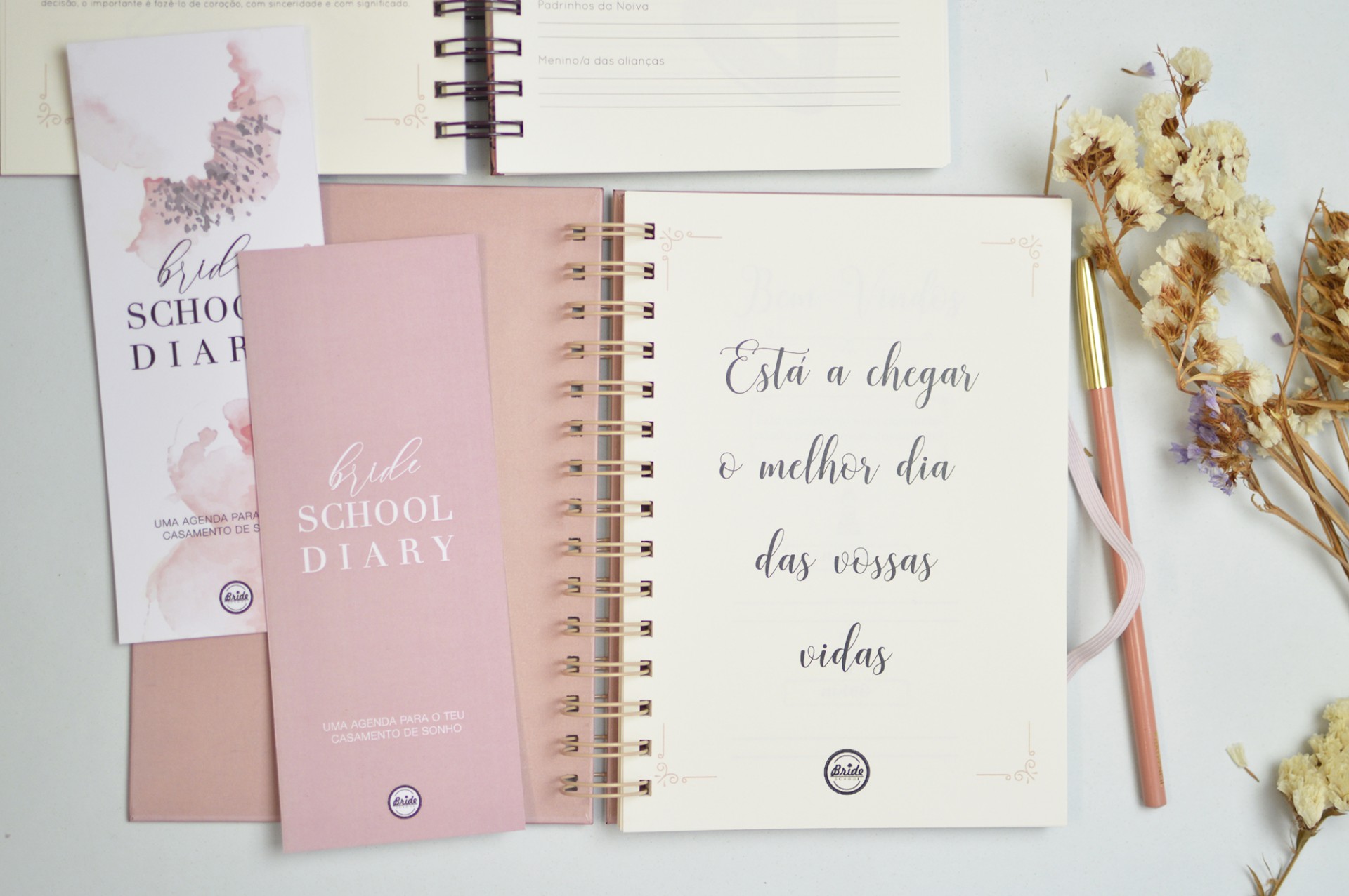 Agenda rosa claro com espiral dourada e texto em português sobre mesa clara com flores secas e folhetos.