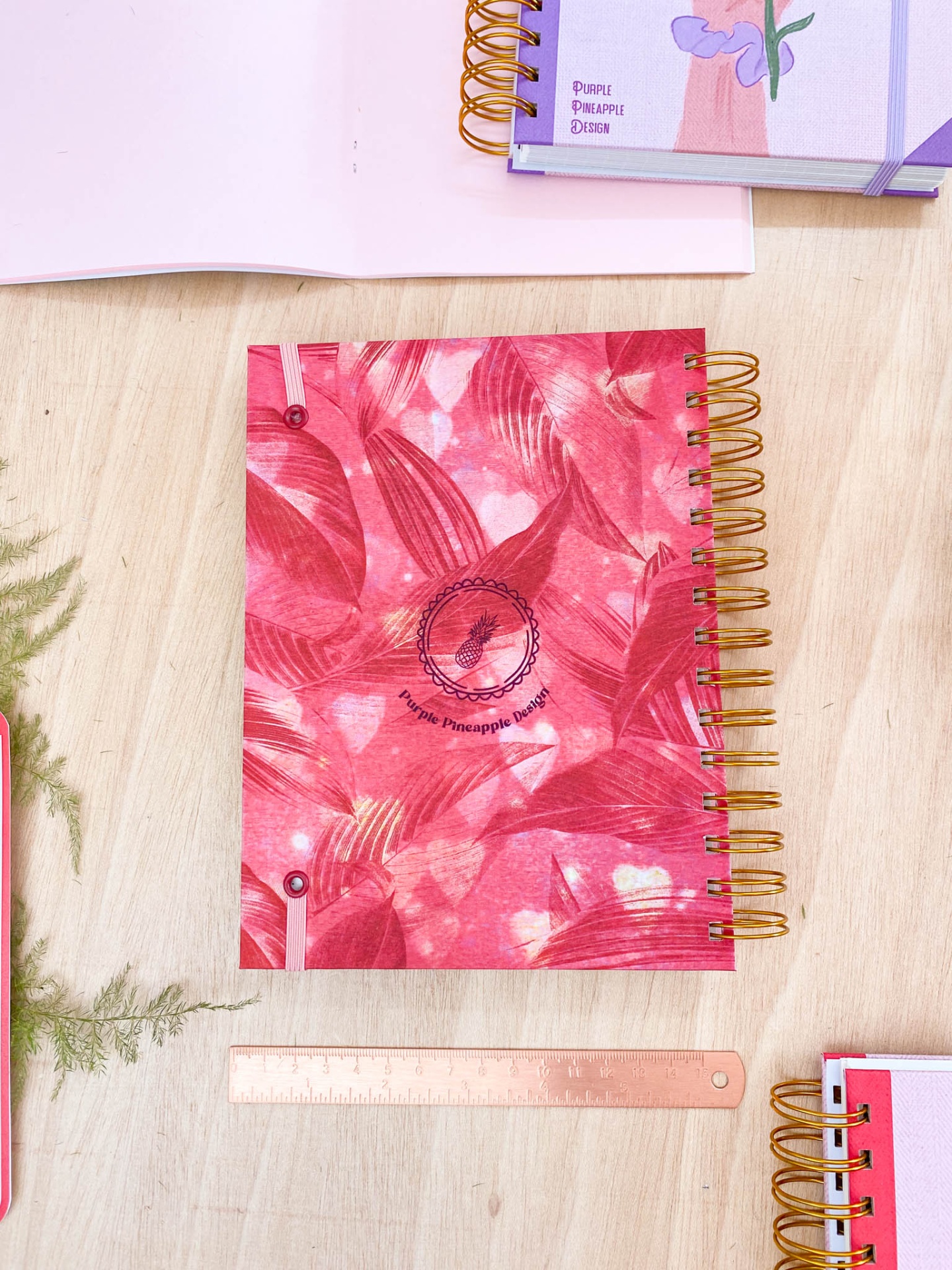 Caderno espiralado vermelho com padrão de folhas e texto Purple Pineapple Design sobre mesa de madeira clara