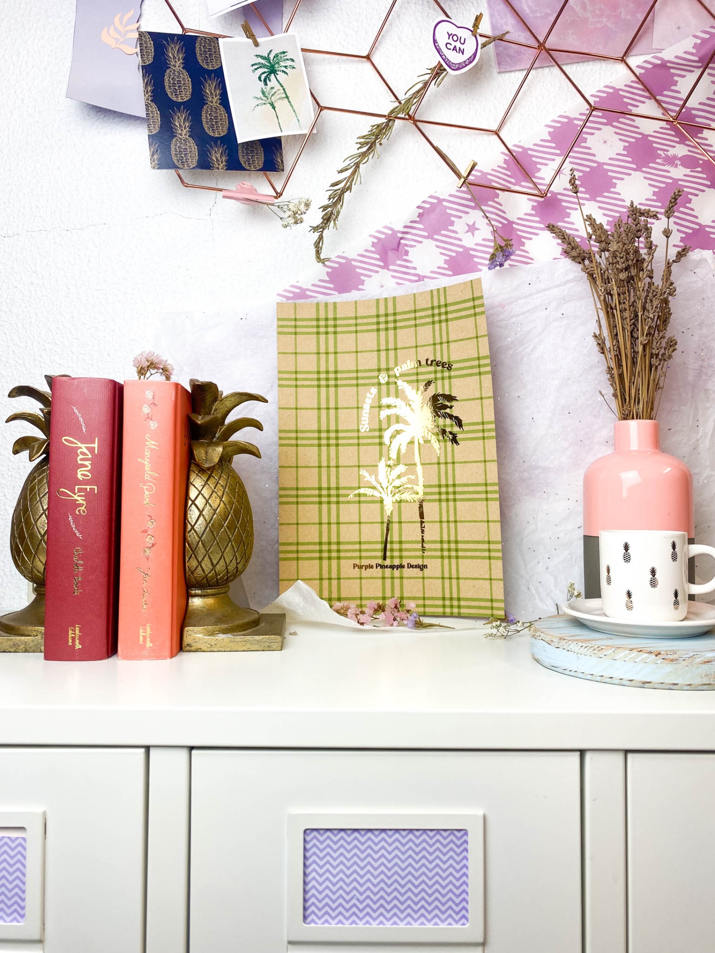 Prateleira com livros, suporte ananás, embalagem xadrez com palmeira e jarra rosa com flores secas