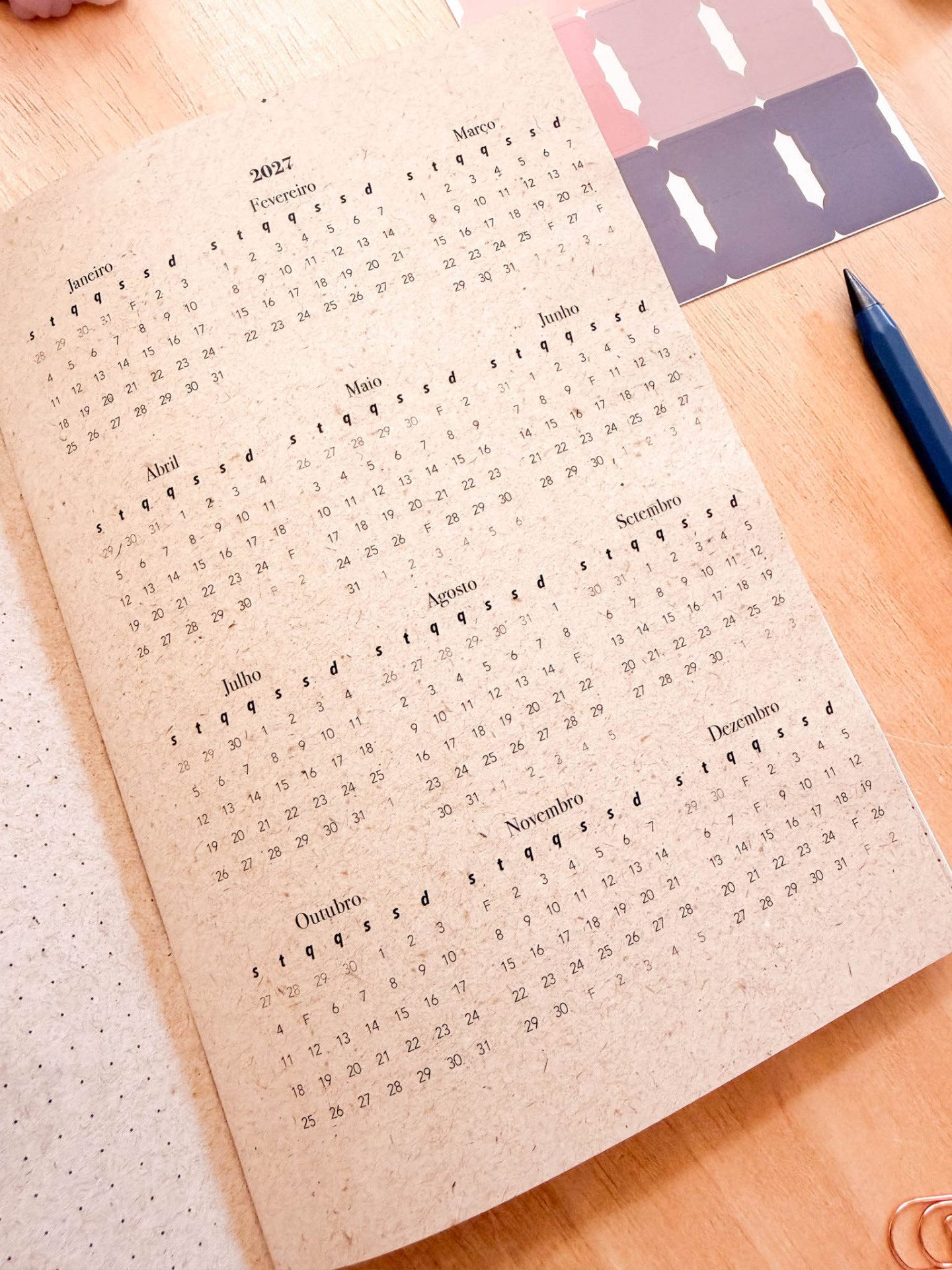 Calendário anual 2027 em papel reciclado sobre mesa de madeira com caneta azul e autocolantes.