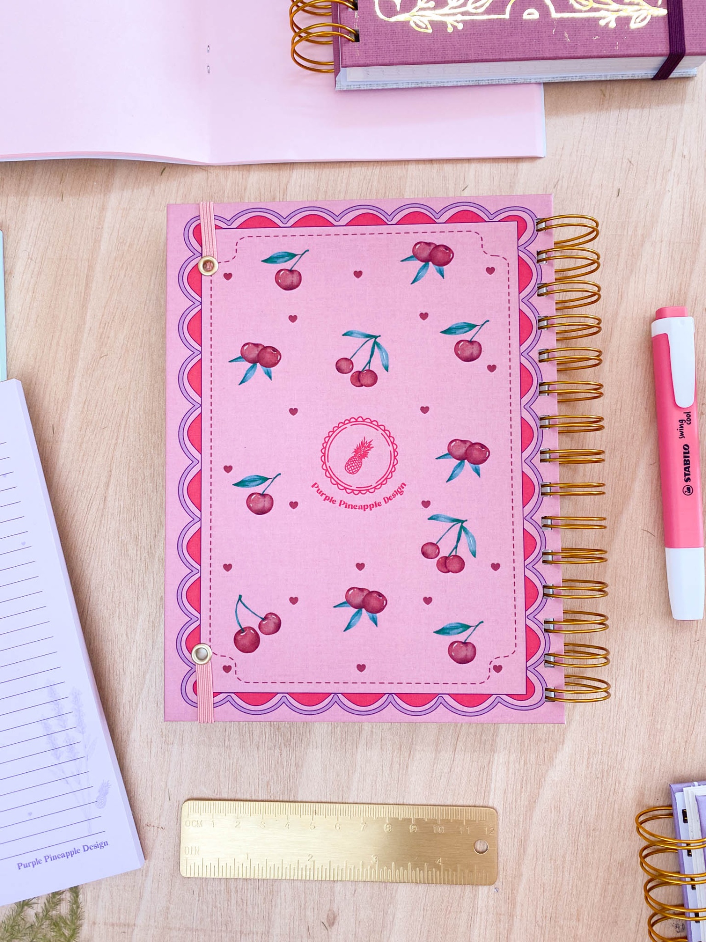 Caderno rosa com padrão de cerejas, argolas douradas e elástico