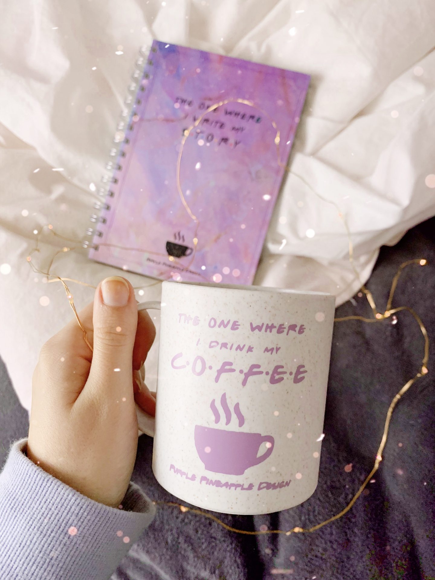 Caneca branca com estampa lilás e texto, mão a segurá-la, caderno lilás de fundo em tecido branco e cinzento.