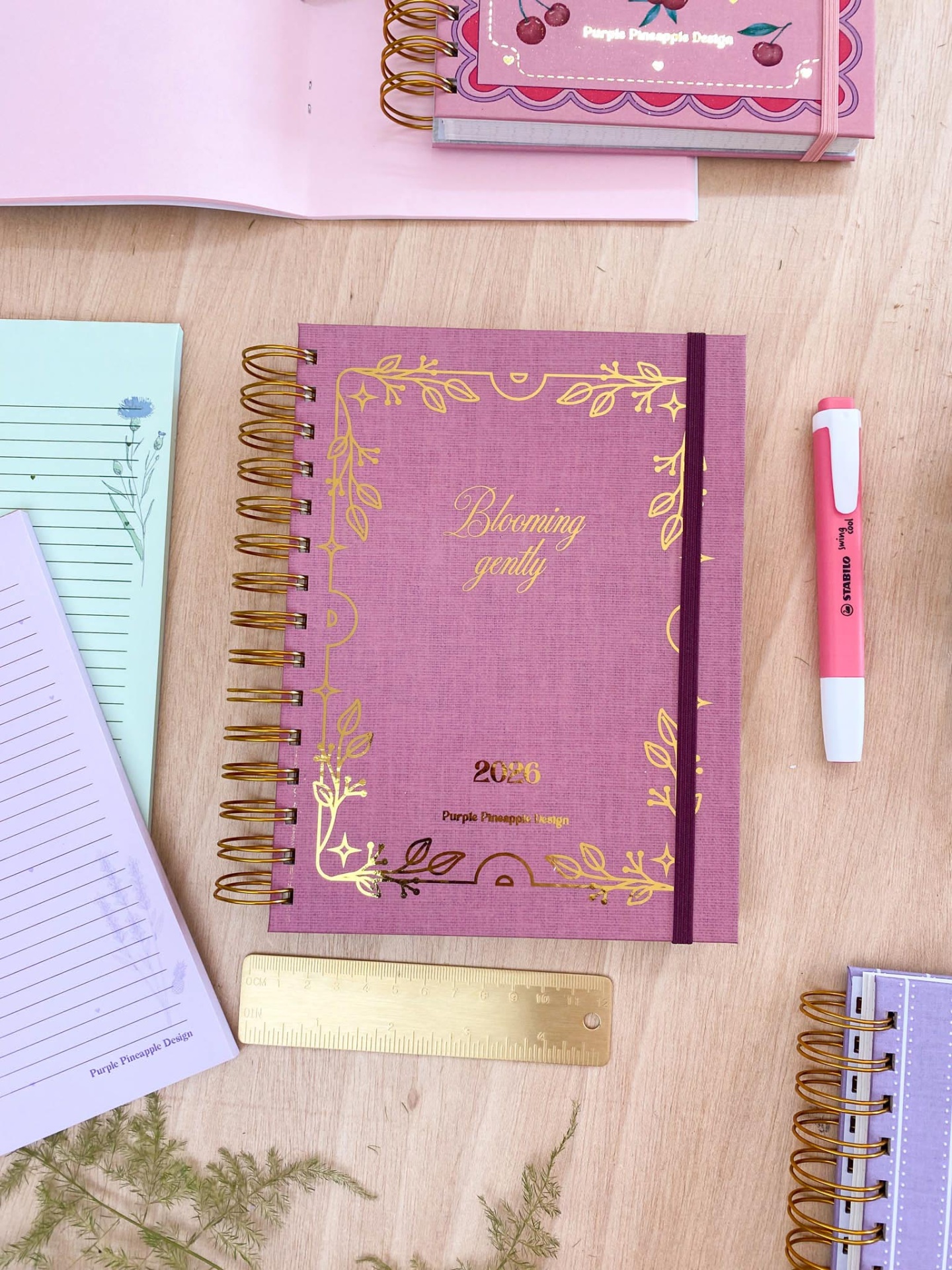 Caderno rosa com elástico roxo e texto dourado Blooming gently em mesa de madeira com caneta e régua