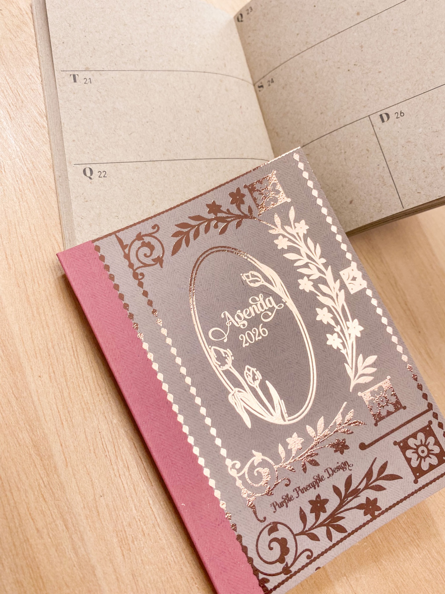 Agenda 2026 com capa cinza e rosa decorada com detalhes dourados sobre mesa de madeira.