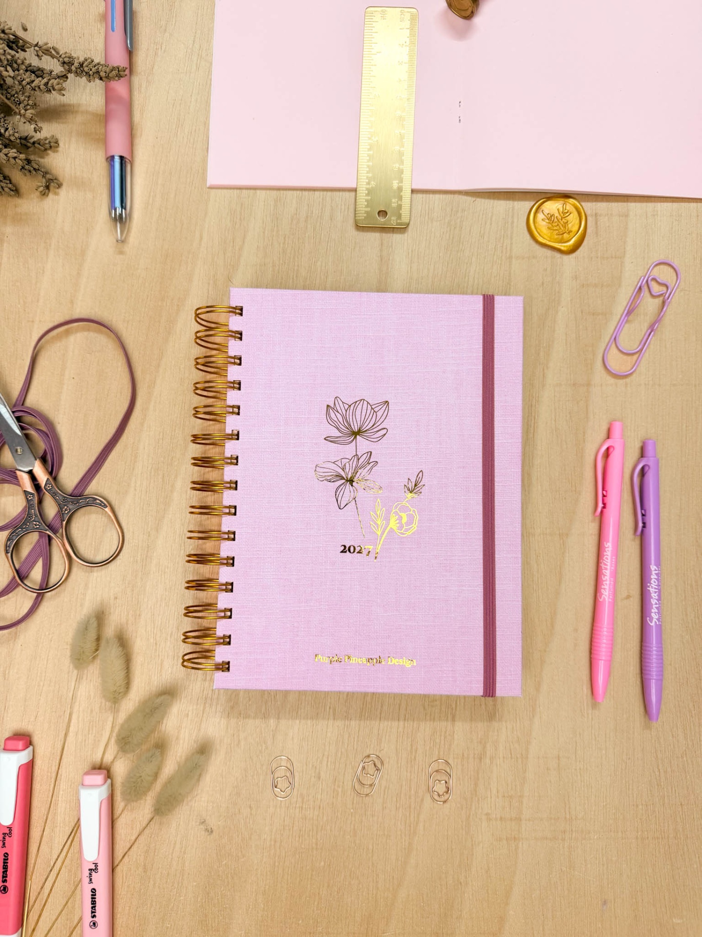 Caderno rosa 2027 com tema floral, tesoura, régua dourada e canetas coloridas numa mesa de madeira