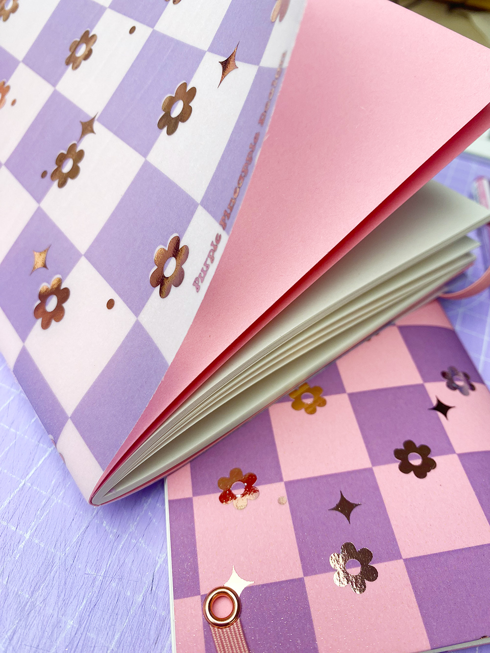 Caderno com capa xadrez lilás e branco com flores douradas, páginas brancas e contracapa rosa