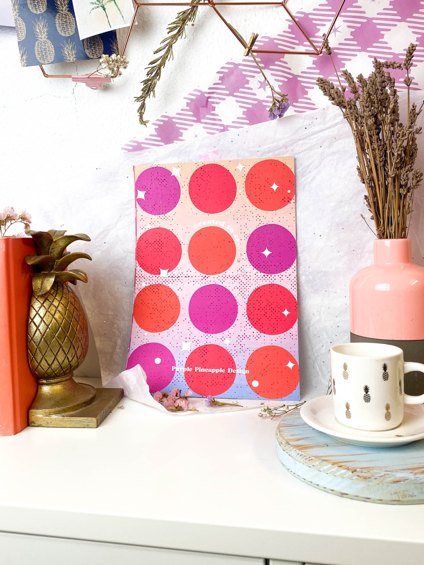 Mesa branca com livros, decoração de ananás dourado, caderno cor-de-rosa com círculos, jarra rosa com flores secas e chávena branca com padrão de ananás