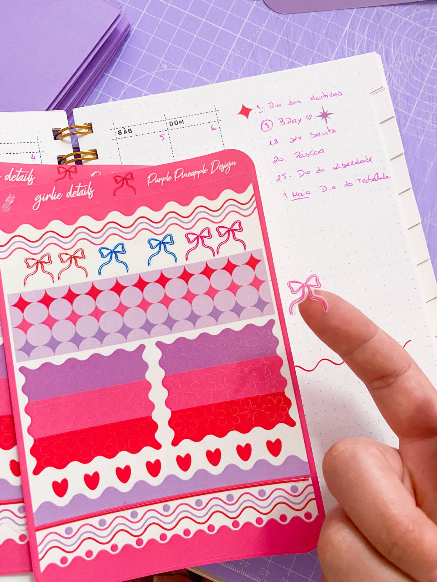 Folha de autocolantes coloridos em rosa, vermelho e roxo com padrões variados junto a caderno aberto com texto em tinta cor-de-rosa.