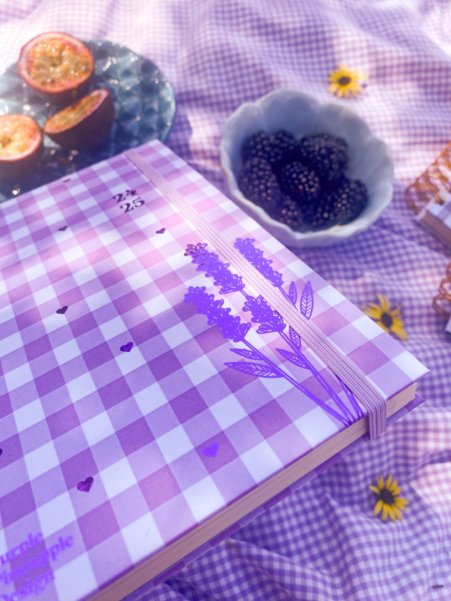 Caderno lilás xadrez com flores ao lado de maracujás e amoras