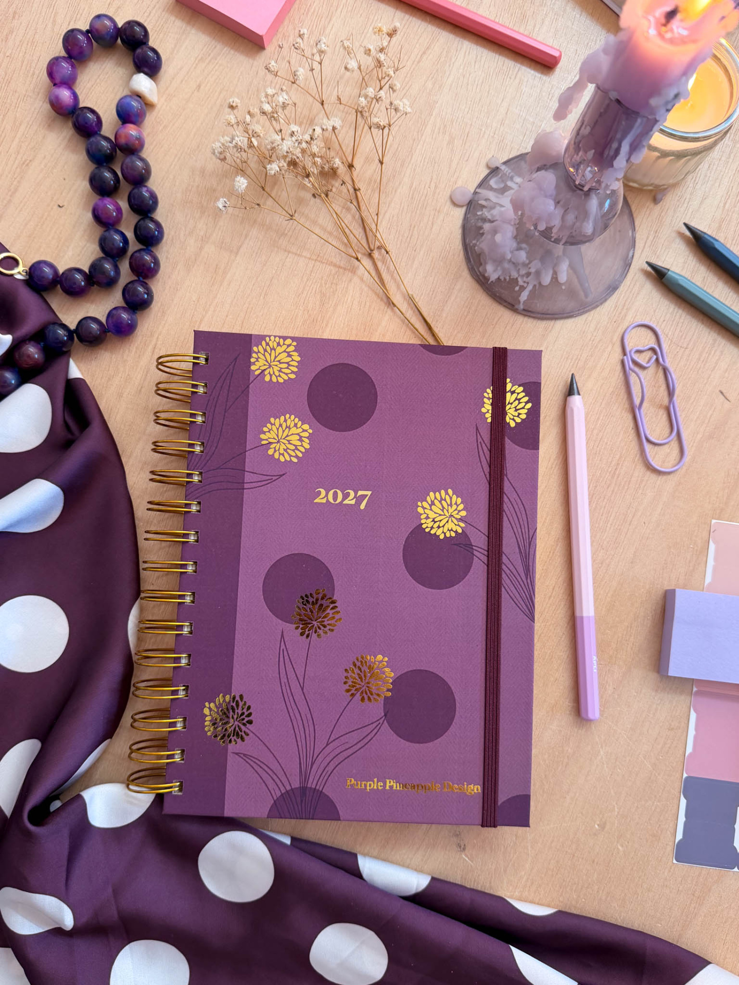 Agenda roxa 2027 com flores douradas, colar de contas, vela derretida e utensílios de escrita numa mesa de madeira.