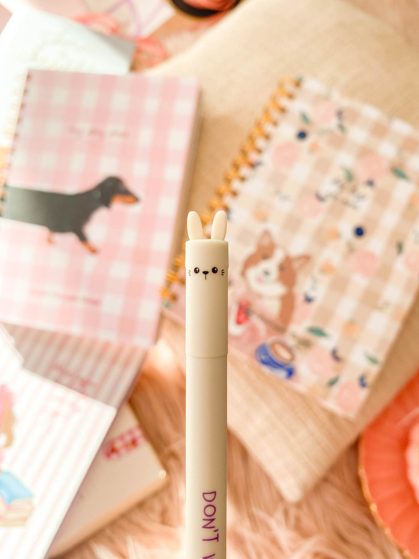 Caneta branca com tampa de coelho e texto roxo, agendas com padrões e cães ao fundo