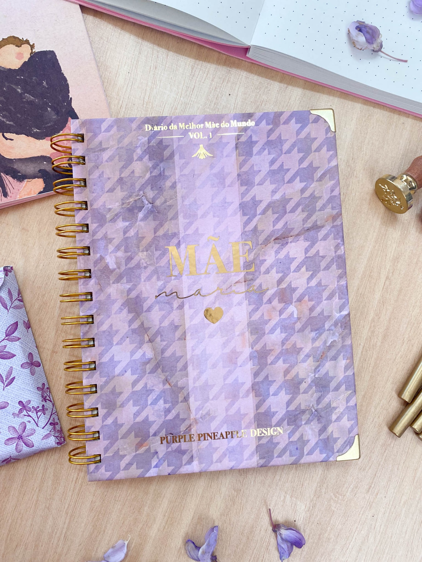 Caderno lilás com padrão pied-de-poule e texto dourado sobre uma mesa de madeira clara