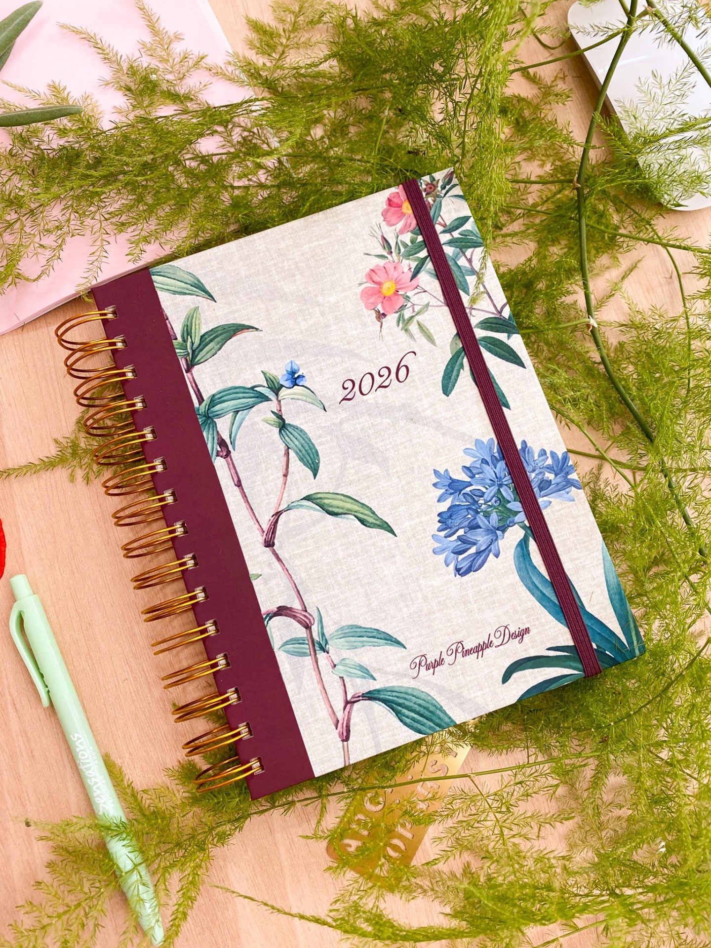 Caderno floral branco com elástico vinho e espiral dourada, texto 2026, sobre mesa de madeira com folhas verdes