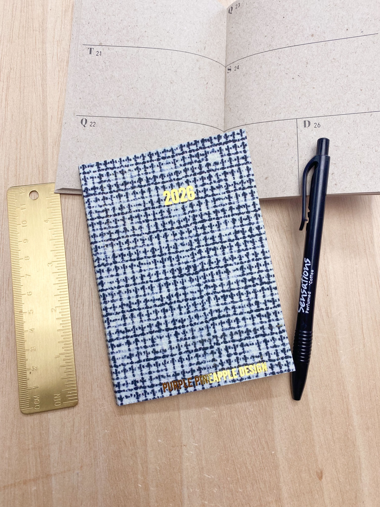 Bloco de notas azul e branco com texto dourado, caneta preta e régua dourada sobre mesa de madeira clara