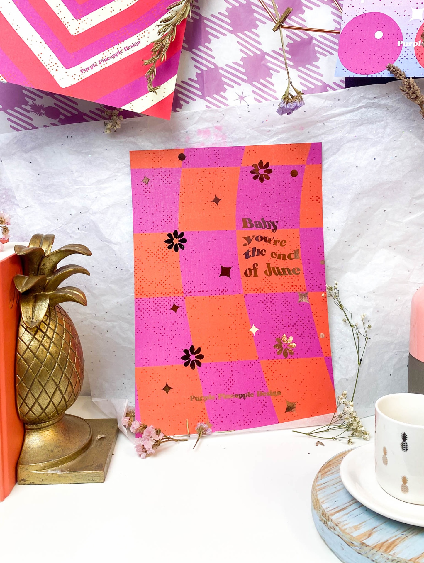 Cartaz decorativo rosa e laranja com texto dourado junto a suporte de abacaxi dourado, livro laranja e chávena