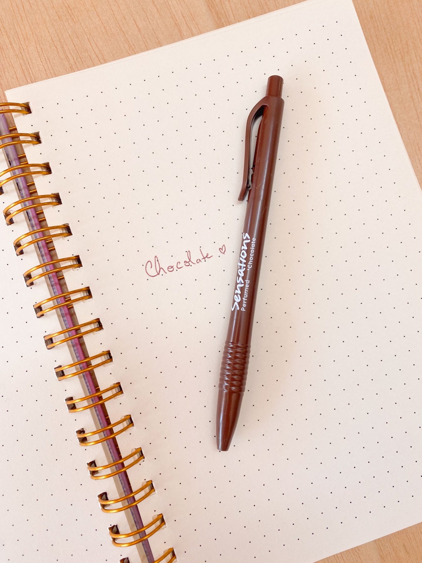 Caderno bege com folhas pontilhadas e caneta castanha com escrita 'Chocolate ♥'