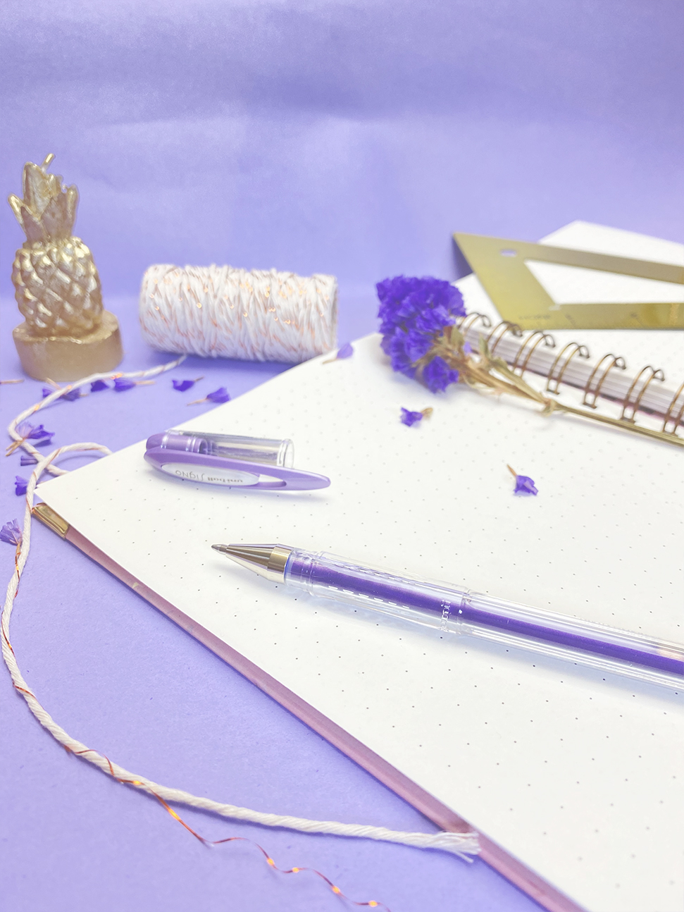 Bloco de notas com caneta, flores secas, régua e vela decorativa sobre fundo lilás