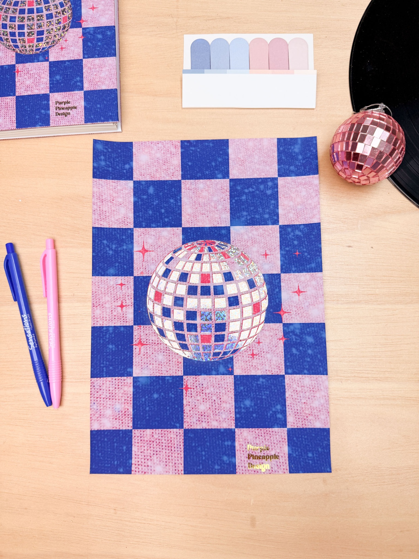 Folha de papel azul e rosa com esfera disco espelhada e vários objetos de papelaria numa mesa de madeira